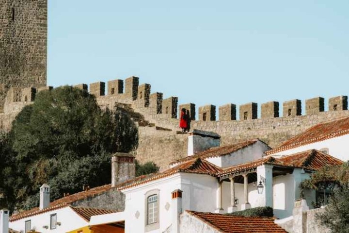 Stone castle wall with person in red above terracotta-roofed buildings.